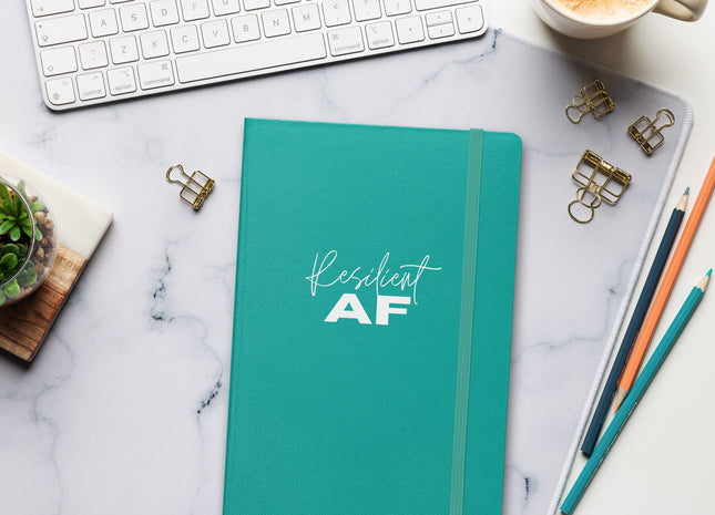 A blue hardcover notebook with the text 'Resilient AF' written on the cover, placed on a desk alongside a computer, a coffee cup, and some pens.