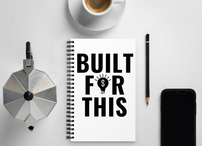 A spiral notebook with a white cover featuring the text 'BUILT FOR THIS' in bold black letters, placed on a desk alongside a coffee cup and a mobile phone.
